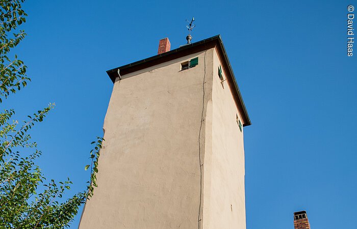 Blick von unten auf einen hohen Turm mit beiger Fassade. Daneben ein Abschnitt eines Hauses mit gleicher Fassade, einem Fenster und grünem Fensterladen.