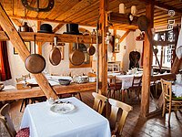 Eine Raum getrennt durch Holzbalken. Im raum stehen Tische mit weißen Tischdecken. Auf den Balken stehen alte Töpfe, Kerzen, Backformen und Hüte.