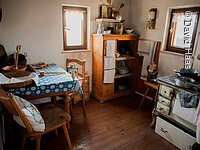 Eine kleine altertümliche Küche. Sitzecke mit zwei Stühlen in der rechten Ecke. in der hinteren Ecke ein Holzschrank. Rechts davon ein alter Holzofen zum Kochen.