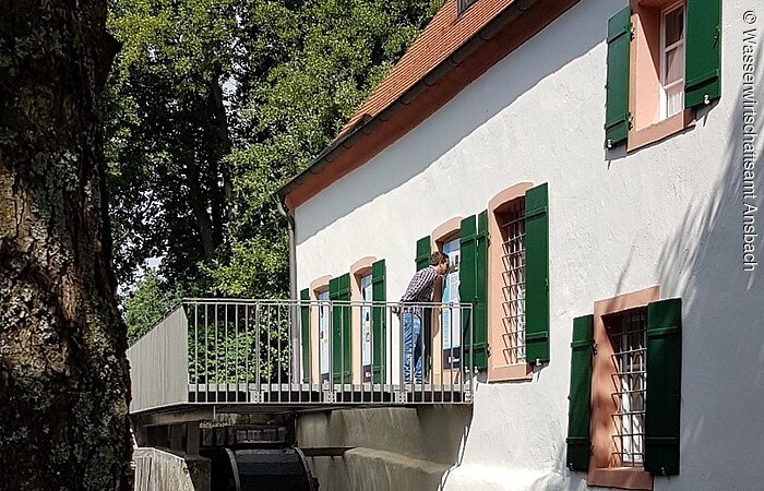 Einblicke in den ehemaligen Mahlraum der Mandlesmühle Mandlesmühle von außen. Ein großes weißes Haus mit grünen Fensterläden. Entlang des Hauses fließt ein kleiner Bach.