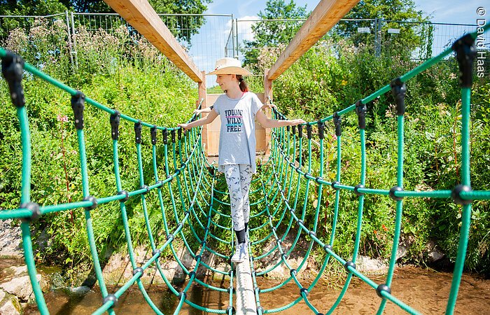 Ein Mädchen läuft über einen Balken. Links und rechts ein grünes Netz zur Sicherung. Darunter fließt ein Bach.