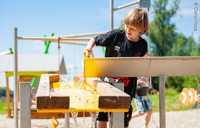 Ein junge steht auf einem Spielplatz und spielt mit Wasser. Er kippt Wasser in einen gelben Tisch.