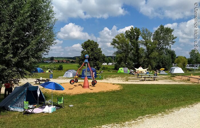 Ein Spielplatz mit kleinem Karusell. Rechts davon eine Tischtennisplatte.Im Hintergrund stehen Zelte auf einer grünen Wiese.