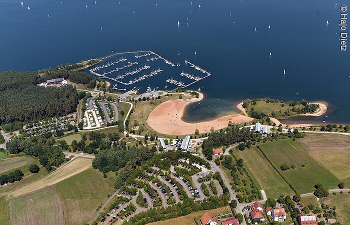 Luftbild vom Seezentrum Ramsberg. Grüne Felder und Wiesen, es folgt ein Snadstrand direkt am Ufer des Brombachsee. Im See ein Segelhafen.