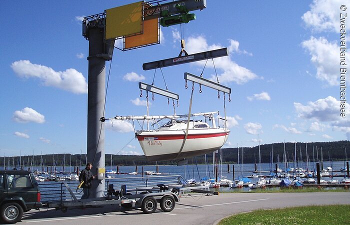 Ein Segelboot wird mit der Hilfe Bootskran auf den Anhänger eines Autos geladen. Das rot-weiße Boot hängt in der Luft. Ein Mann bedient den Kran.