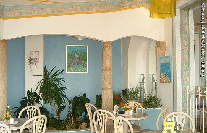 Ein Gastraum mit türkis weißen Wänden. Im Gastraum stehen drei runde Tische mit weißen Stühlen.