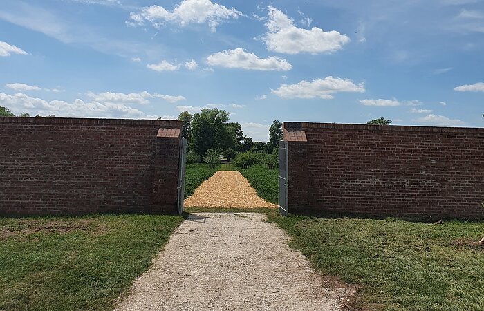 Schotterweg der auf ein Tor in einer roten Ziegelsteinmauer zuführt.