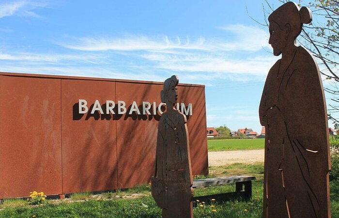 Zwei Frauenfiguren aus Metall ausgeschnitten stehen auf einer Wiese. Dahinter ein Metallbau mit dem Wort Barbaricum.