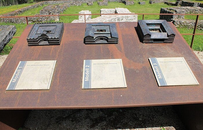 Auf einer Metallplatte sind drei verschiednee Modelle von römischen Bauwerken befestigt. Unter jedem Modell ist eine Informationstafel angebracht.