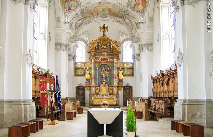 Innenansicht der St. Nikolauskirche. Altarraum mit Holzbänken entlang der Wände. Hinten in der Mitte ein großer prunkvoller Altar aus Gold.