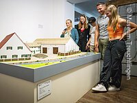 Nachbildung Jura-Bauernhof Besucher Vier Gäste in der Abteilung Möbel aus dem 17. Jahrhundert. Sie stehen auf der rechten Seite und blicken auf das Modell eines Jura-Bauernhofes.