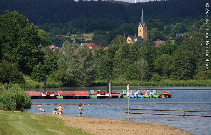 Blick auf den Brombachsee in Pleinfeld. Im Vordergrund sind badende Kinder am Strand, dahinter Trettboote am Liegeplatz zu erkennen. Im Hintergund ragt die Pleinfelder Kirche zwischen Bäumen hervor.