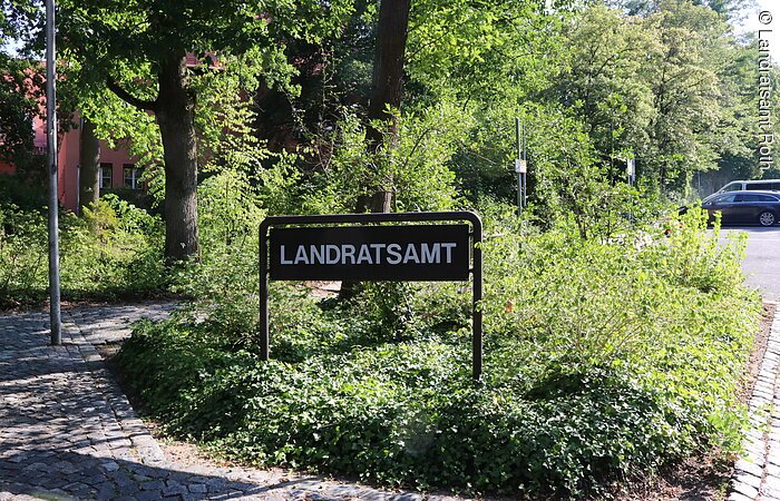 Schild mit dem Wort Landratsamt. Das Schild steht in einer kleinen Grünfläche mit Büschen. Links verläuft ein Fußweg. Rechts davon eine Straße.