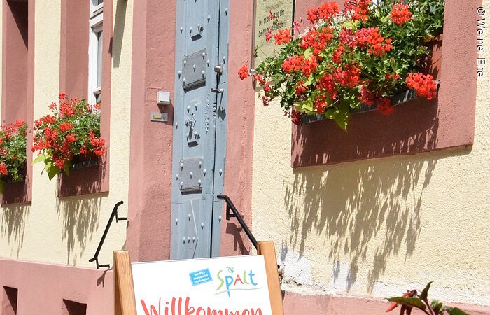 Stadtbücherei Spalt Eingang eines Hauses im fränkischen Baustil. gelbe FAssade mit roten Akzenten. Eine große graue Eingangstür aus Metall. Davor ein Schild mit dem Wort Willkommen und einem Foto eines Gartens mit grünem Gewächs.