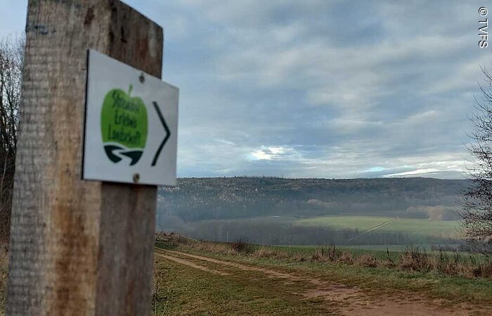 Berglandschaft Blick auf einen grünen Berg in der herbstlicher Landschaft. Im linken Bildrand ein Holzposten mit einem Schild. Ein grüner Apfel auf weißem Grund mit der Aufschrift Streuobst Erlebnislandschaft.