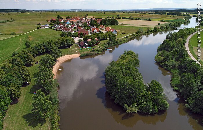 Ein Luftbild des Zuleiters in Ornbau. Am Ufer wachsen grüne Bäume auf grünen Wiesen. Im hinteren Bildabschnitt sieht man die Häuser eines kleinen Dorfes.