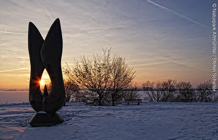 Kunstobjekt bei Sonnenaufgang am schneebedecktem Altmühlsee. Einige Meter dahinter stehen einige Sträucher.