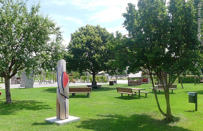Eine große Rasenfläche mit Sitzgruppen aus Holz. Auf dem Platz zwei Kunstobjekte geschnitzt aus Holz. auf der Wiese stehen außerdem Bäume. Im Hintergund erkennt men die Spielgeräte eines Spielplatzes.