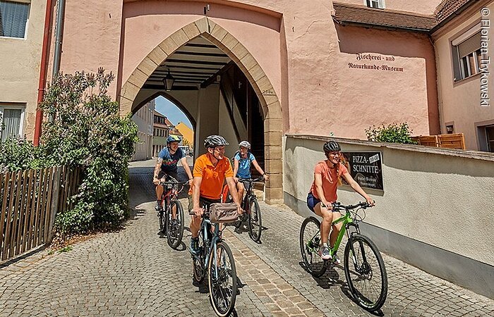 Vier Radfahrer radeln über eine geplasterte Straße durch ein Stadttor. Das Stadttor hat eine rosane Fassade und blau-weiß gestreifte Fensterläden.