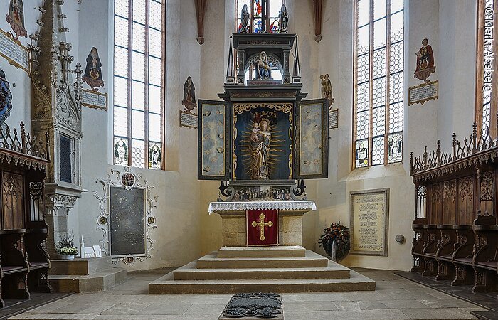 Innenraum einer Kirche. Weiße Fassade mit hohen Fenstern. Blick auf den Altar mit Treppe und Jungrau Maria mit Kleinkind auf dem Arm. Links und rechts vom Altar Bänke aus dunklem Holz.
