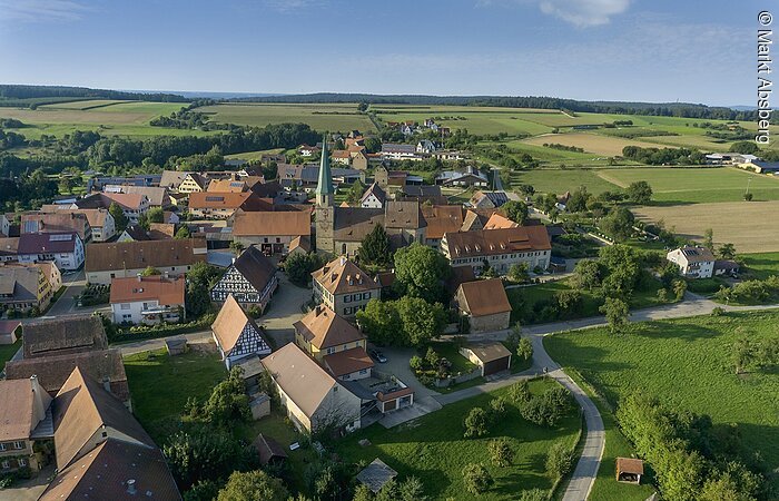 Luftbild von Kalbensteinberg. Blick auf die Rieter-Kirche umgeben von Wohnhäusern. Um den Ortsteil erstrecken sich grüne Felder.