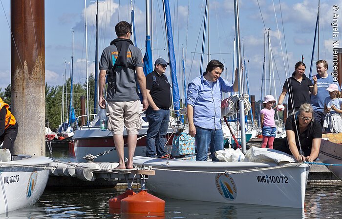 Urlauber stehen auf Steg eines Segelboothafens. Eine Frau vernkoten die Seile eines Segelboot. Ein Mann schaut ihr dabei zu. Links von ihm steht ein Mann mit Schwimmweste mit dem Rücken zur Kamera. Dahinter stehen noch vier weiter Urlauber am Steg.