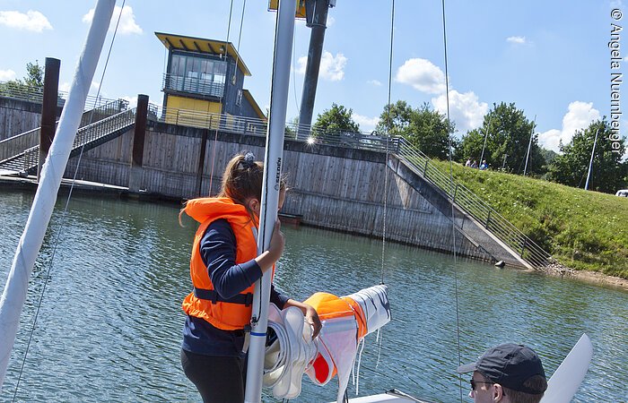 Zwei Segler mit Schwimmweste bereiten ihr Segelschiff für eine Ausfahrt vor. Dabei hilft ihnen eine Mitarbeiterin der Segelschule Fürst. Eine Frau steht auf dem Segelschiff, dass an einem Holzsteg im Wasser liegt. Die zwei anderen halten es fest. Die Sonne scheint.