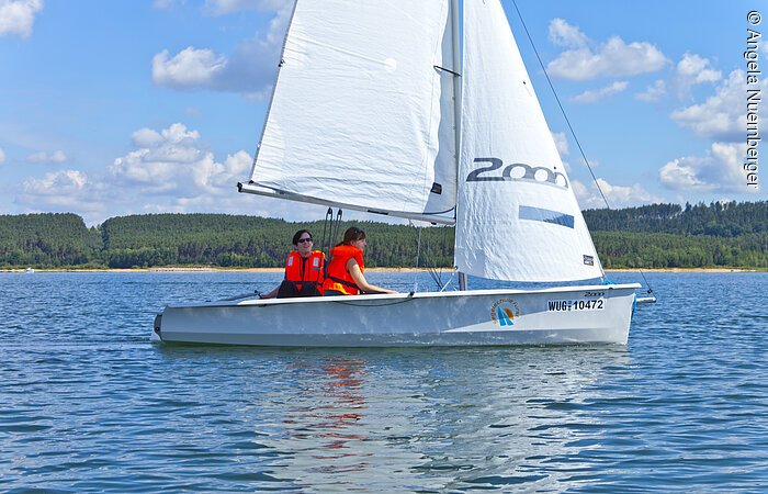 Zwei Segler mit oranger Warnweste fahren mit ihrem weißen Segelboot über den See. Am Ufer im Hintergund erstreckt sich ein grüner Wald unter blauem Himmel.