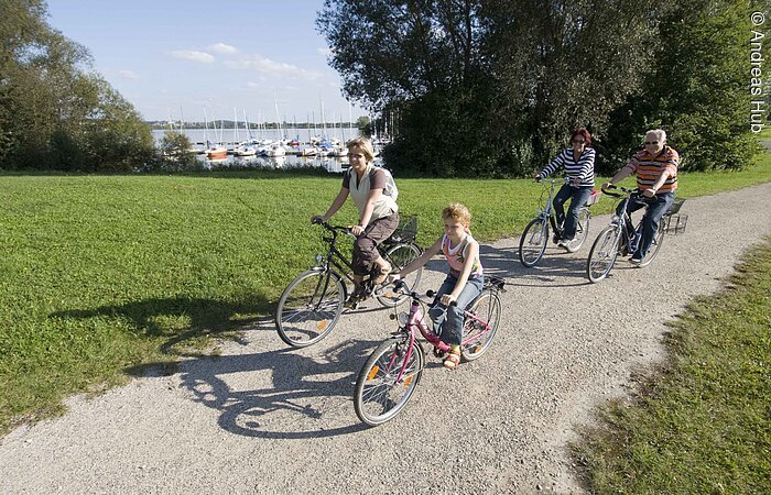 Radtour um den Altmühlsee Drei Erwachsene und ein Kind fahren mit ihren Fahrrädern auf einem geschotterten Fahrradweg entlang des Altmühlsee. Um den Weg herum ist grüne Wiese. Im Hintergund hat man zwischen Bäumen und Sträuchern Blick auf den Hafen des Altmühlsees.