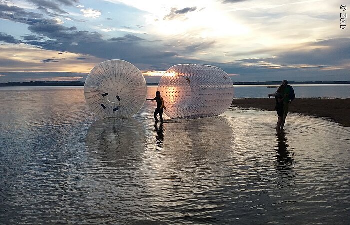 Zwei transparnete doppelhüllige Kugeln schwimmen im Sonnenuntergang auf dem Wasser. Zwei Mädchen stehen dabei. Man sieht nur ihre Silhouetten.