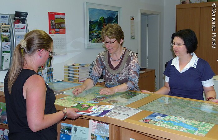 Zwei Frauen stehen hinter einer Beratungstheke einer Touristinformation. Vor der Theke eine Urlauber mit einer Übersichtskarte in der Hand. Eine Frau hinter der Theke spricht mit der Urlauberin und zeigt dabei auf die Karte. Auf der Theke liegen weitere Karten und Infomaterial.