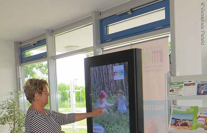 Eine Frau steht vor einer Infosäule und bedient diese. Die Infosäule ist ein stehender Bildschirm mit Touch-Funktion.