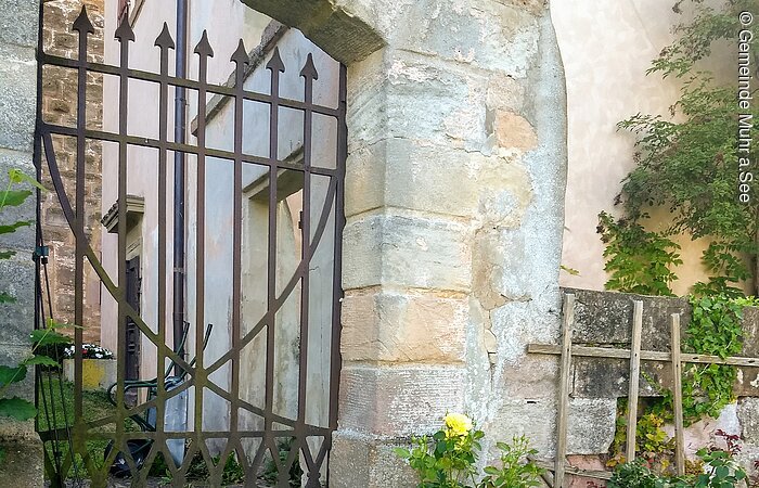 Ein Tor aus Metall in einem Gemauerten Steinbogen. An die Mauer schließt ein Haus an.