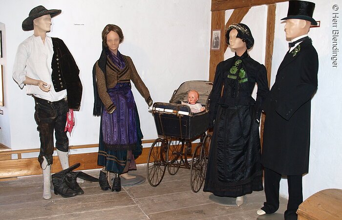 Vier Schaufensterpuppen stehen in einem Raum mit weißer Wand und braunem Fachwerk. Die Puppen tragen fränkische Trachten. In der Mitte ein alter Kinderwagen.