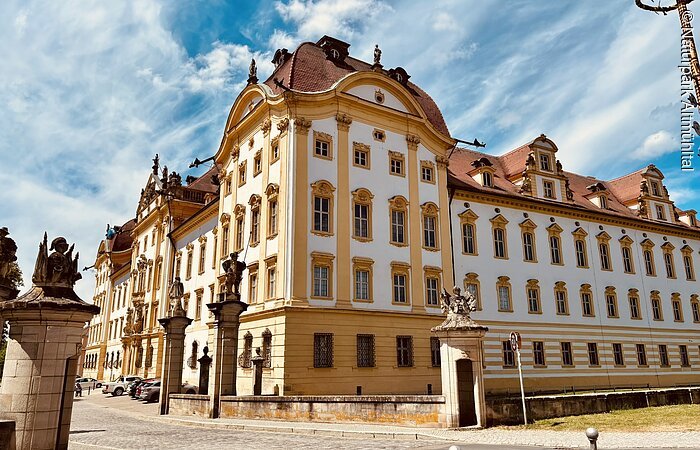 Zentrales Element des Bildes ist die Schlossresidenz Ellingen mit kräftigen Farben vor einem blauen Himmel. Links ist ein Pfeiler mit einer Statue zu sehen