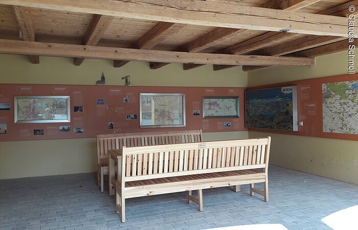 Limes Infopunkt Innenraum eines offenen Häuschen. Darin stehen eine Sitzgruppe aus Holz. An der gelb-roten Wand hängen Informationen zum Limes.