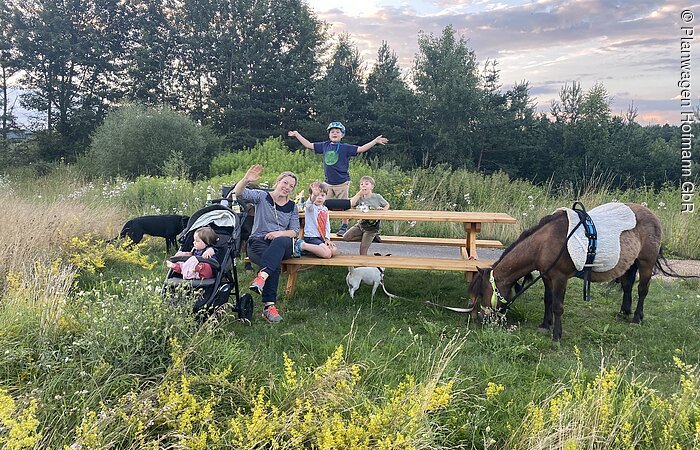 Eine Familie lächelt in die Kamera. Sie sitzen auf einer Holzbank mit Tisch auf einer grünen Wiese. Links steht ein Kinderwagen, rechts ein grasendes Pferd.