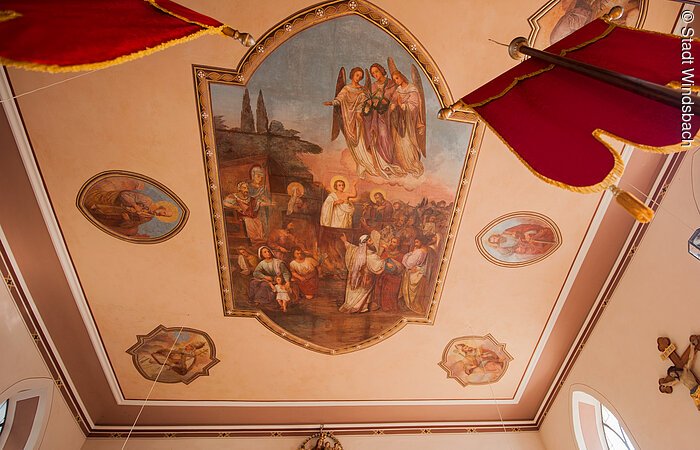 Eine weiße Decke einer katholischen Kirche. Darauf ein Gemälde mit Engeln. Unter ihnen stehen Menschen mit langen Gewändern.