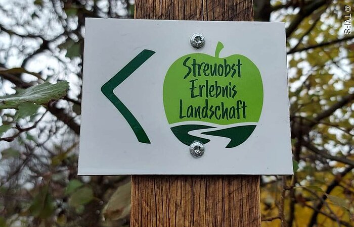Ein Hinweisschild des Streuobst Erlebnislandschaft, ein grüner Apfel auf weißem Hintergund, hängt an einem Holzpfosten.