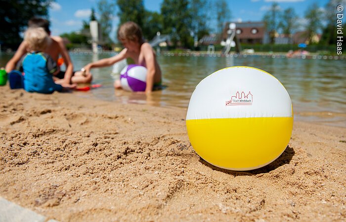 Im Vordergrund ein Wasserball am Strand. Dahinter spielen Kinder am Strandufer.