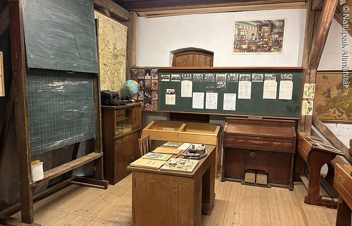 Ein altes Klassenzimmer mit Tafeln, einem Pult und Schulbänken. Das Pult steht in der Mitte.