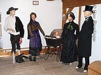 Vier Schaufensterpuppen stehen in einem Raum mit weißer Wand und braunem Fachwerk. Die Puppen tragen fränkische Trachten. In der Mitte ein alter Kinderwagen.