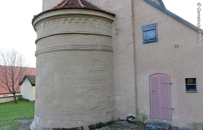 Außenansicht einer Kirche mit heller brauner Fassade. Zu sehen ist ein Wand des Kirchenschiffs mit kleinem runden Turm.