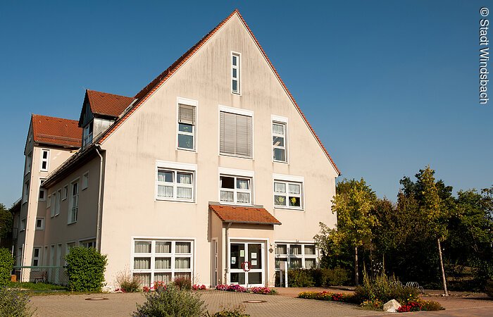 Ein großes Haus mit vier Ebenen. Helle rosane Fassade mit weißen Fenstern. Rechts davon Bäume.