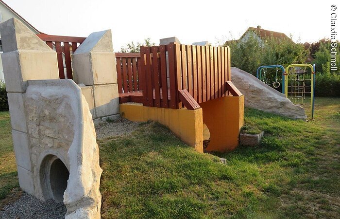 Auf einem Spielplatz steht eine Ausichtsplattform aus Stein. Darunter hindurch führt ein Tunnel durch.