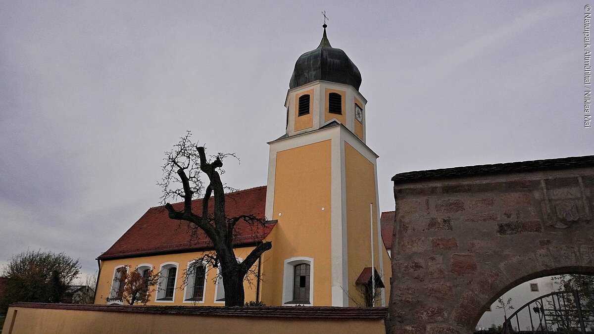 Kirchturm mit Zwiebeldach und gelbes Kirchengebäude hinter einer Mauer bei bewölktem Himmel.