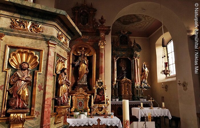 Innenansicht einer Kirche mit Altären, goldverzierten Heiligenfiguren und Kerzen auf Tischen mit weißen Tüchern