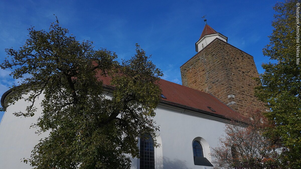 Kirche mit weißer Fassade, rotem Dach und steinernem Turm vor blauem Himmel und Bäumen im Vordergrund.