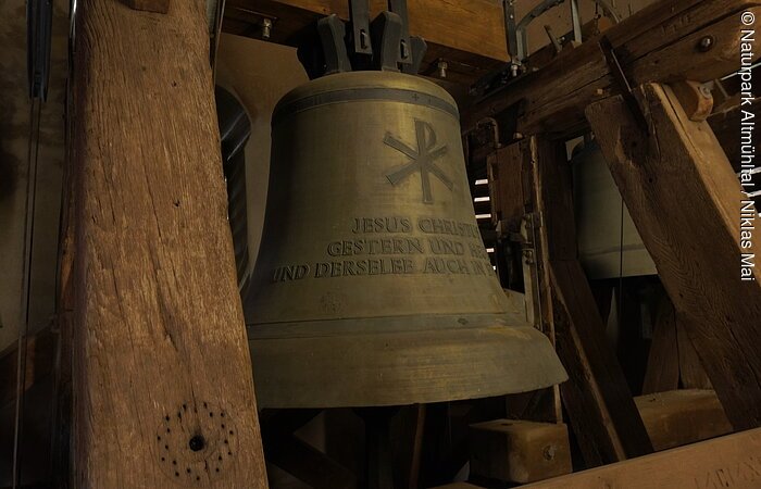 Große bronzene Kirchenglocke mit christlichem Symbol und Schriftzug, umgeben von Holzbalken im Glockenturm.