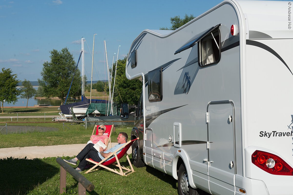 Eine Frau und ein Mann sitzen in zwei Liegestützen link neben einem Wohnmobil. Im Hintergund lagern Boote auf Anhängern. Dahinter beginnt der See.